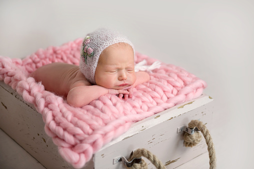 Sleeping newborn baby lying in box on pink rug with hands folded together. Newborn little girl with chubby cheeks sleeping in wooden box