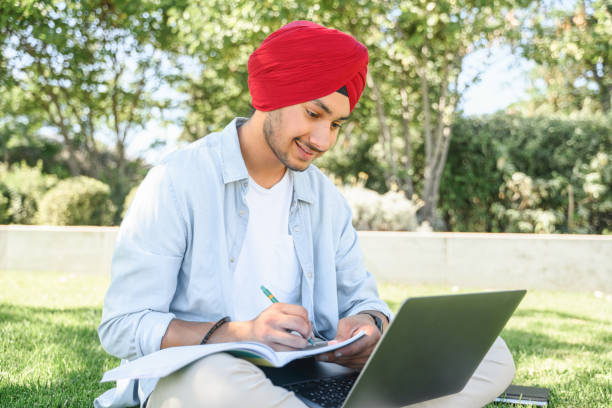 E-learning. Multiracial Indian man in turban using laptop for watching online courses, webinars E-learning. Multiracial Indian man in turban using laptop for watching online courses, webinars, takes notes sitting in the park in campus, male student preparing exams, taking educational classes anhui province stock pictures, royalty-free photos & images