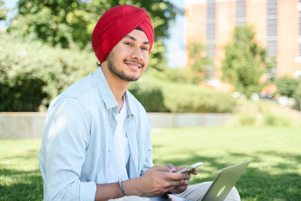 uśmiechnięty indyjski student w narodowym paggu w turbanie siedzący na zewnątrz z laptopem, za pomocą smartfona - technology typing male beauty asian and indian ethnicities zdjęcia i obrazy z banku zdjęć