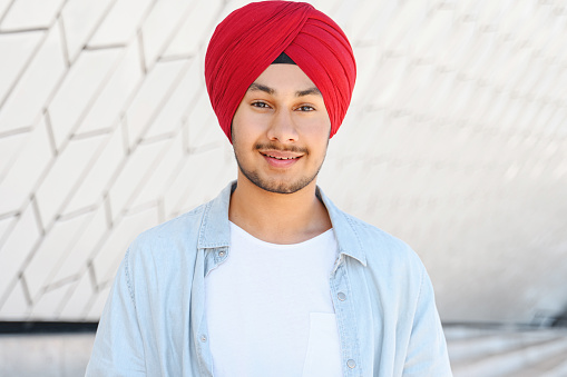 Young handsome Indian man in red traditional turban and casual shirt standing outdoors, smiling pleasant hindu student looking at the camera, headshot