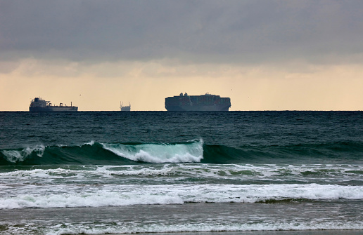 Container ships sitting on the horizon off the Southern California coast with a wave breaking in the foreground