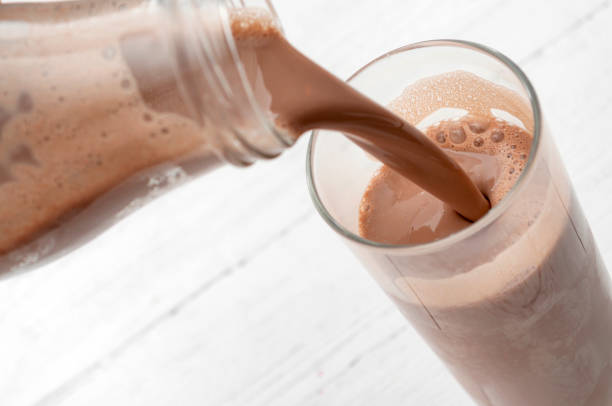 Calcium and protein rich beverage, nutritious milkshake and refreshing treat concept with photograph of bottle pouring chocolate milk into a tall glass isolated on rustic white wood background Calcium and protein rich beverage, nutritious milkshake and refreshing treat concept with photograph of bottle pouring chocolate milk into a tall glass isolated on rustic white wood background protein drink stock pictures, royalty-free photos & images