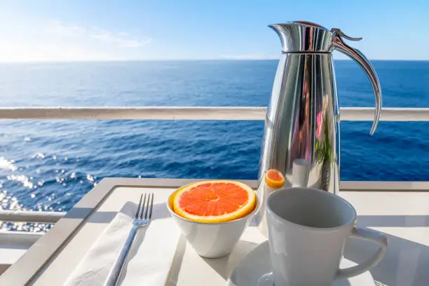 Photo of A carafe of coffee with a coffee cup alongside a cut grapefruit on a ocean view balcony of a cruise ship at sea on a summer day.