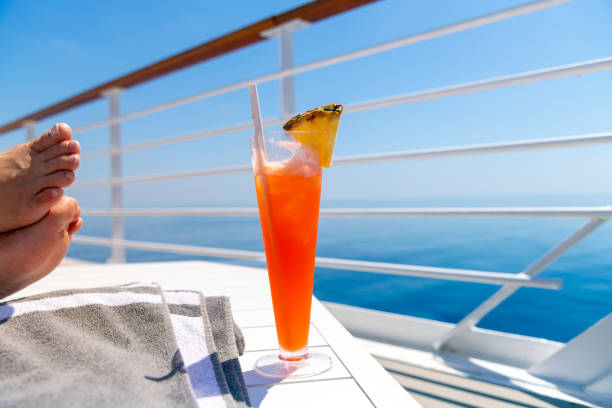 una donna appoggia i piedi su un tavolo sul ponte superiore di una nave da crociera in mare con una bevanda colorata con ananas in una calda giornata estiva sull'oceano. - nave da crociera foto e immagini stock