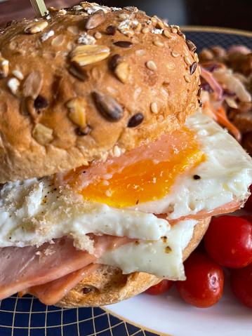 Stock photo showing close-up view of blue rimmed plate with homemade breakfast roll filled with slices of ham and fried eggs with runny yolks served with cherry tomatoes.