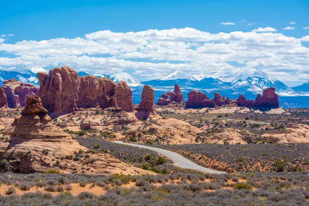 A winding road runs through the Arches National Park. Behind the dry desert there are snow capped mountains. Seen at day in the summer.