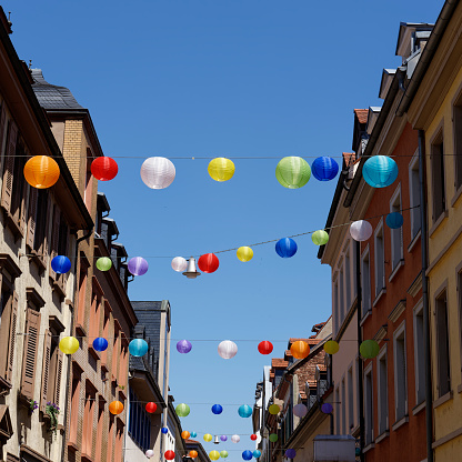 Many colorful lanterns in the sky between historic houses under a bright blue sky.