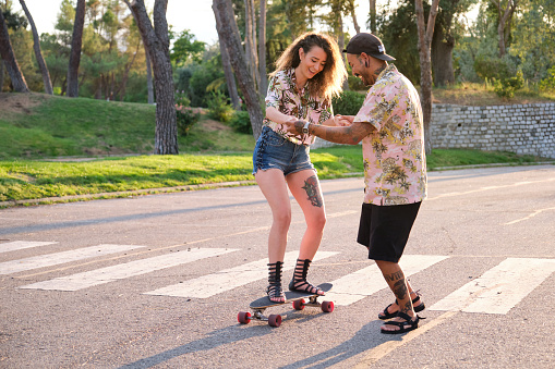Young cool tattooed man teaching his girfriend how to skateboarding on a road in a park.