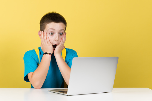 Surprised boy hands on face in front of computer isolated on yellow background.