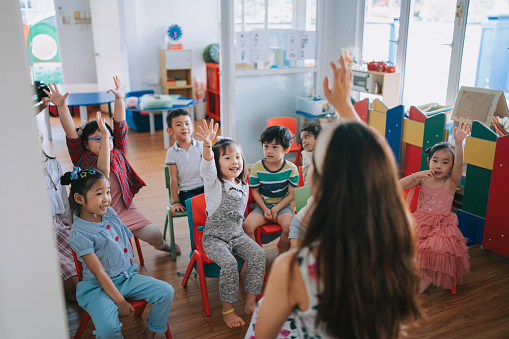 A small group of Kindergarten children sit on the floor of their classroom as their teacher lead them in a sing-along.  They are each dressed comfortably and are smiling as they do the actions to the song.