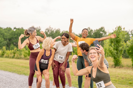 A large group of mature adults casually pose together for a selfie after a successful outdoor running race.  They are each dressed comfortably in athletic wear and have numbers pinned to the front of them as a teammate holds out a cell phone and snaps a photo.