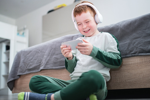 Cute joyful red-haired boy sitting on the floor at the bed and playing games on the smartphone