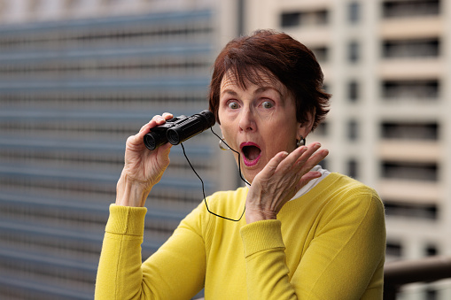 Senior woman holding binoculars looking shocked. Office building exterior in the background.