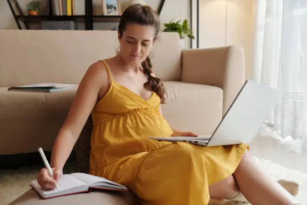 Young pregnant woman sitting on floor with laptop and searching for infor online making notes in her notebook