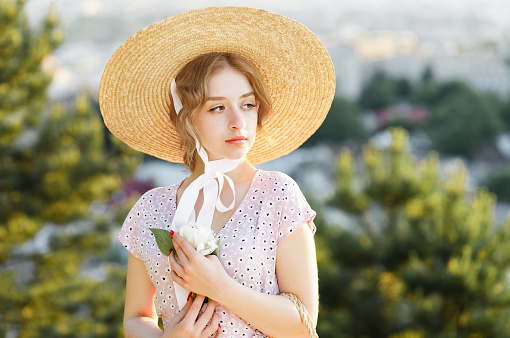 Close-up portrait of a tender blonde in a straw hat posing against the background of the city with a flower in her hand