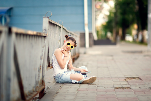 pretty girl with headphones and a phone sits on the sidewalk, a lonely child on a city street. Walking, adolescence and communication difficulties