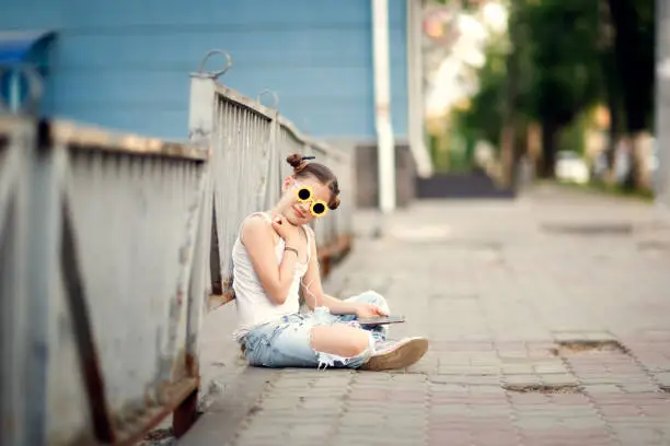 Photo of pretty girl with headphones and a phone sits on the sidewalk, a lonely child on a city street. Walking, adolescence and communication difficulties
