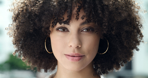 Portrait of a beautiful woman's face with afro curly hair and brown eyes. One stylish and confident mixed race female with cool hairstyle, trendy makeup and hoop earrings standing outside in the city