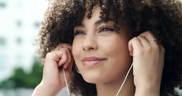 Photo of Closeup of one happy young woman with curly hair listening to favourite music songs or podcast with earphones in the city outdoors. Face of female having fun while exploring and travelling in town