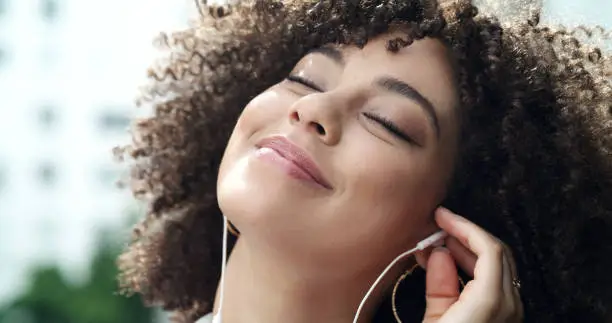 Photo of An edgy lady with an afro listening to music. Glamorous woman with a beautiful face wearing earphones and enjoying her time while listening to a quality playlist. Trendy woman playing a favorite song