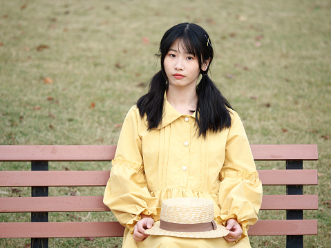 Portrait of Chinese young girl with black long hair in yellow dress sitting and looking at camera on park bench, front view.