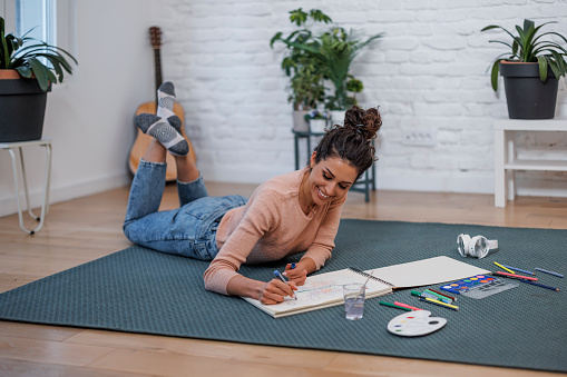A young Caucasian woman is relaxing on her living room floor, painting with watercolors while smiling wide.