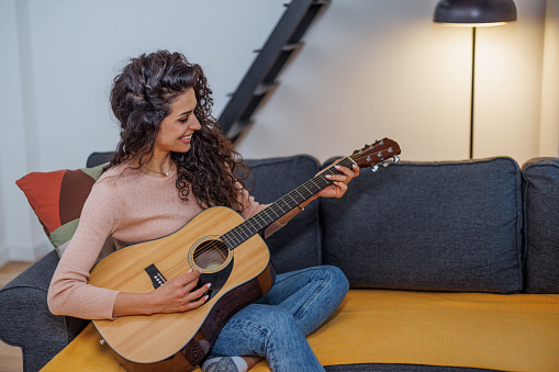 A young Caucasian woman is cheerfully playing the guitar while sitting on her sofa.