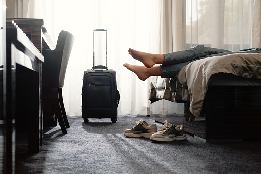 woman taking off footwear in a hotel room on the bed. Tourist relaxing on hotel room after travelling with suitcase. Female having rest after long trip with language Dark silhouette