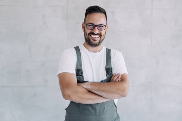 Young male worker in an overall uniform posing at his workplace. Shot of confident smiling professional handyman in overalls and white t-shirt standing with crossed arms and looking at camera. bib overalls stock pictures, royalty-free photos & images
