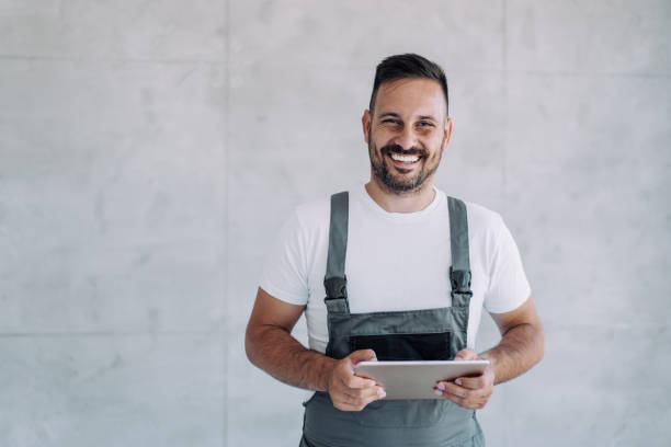 Young male worker in an overall uniform using tablet at his workplace. Shot of confident smiling professional handyman in overalls and white t-shirt using digital tablet. repairman stock pictures, royalty-free photos & images