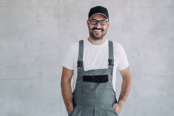 Young male worker in an overall uniform posing at his workplace. Shot of confident smiling professional handyman in overalls and white t-shirt standing with hands in pockets and looking at camera. repairperson occupation stock pictures, royalty-free photos & images