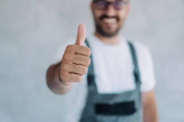 Shot of confident smiling professional handyman in overalls and white t-shirt standing in his workplace and showing thumb up. Focus is on the thumb.