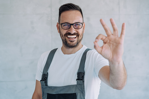 Shot of confident smiling professional handyman in overalls and white t-shirt standing in his workplace and making OK sign with fingers.