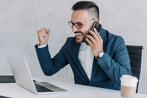 Shot of happy businessman celebrating successful deal in his company. Cheerful businessman talking on mobile phone while using laptop in the office.