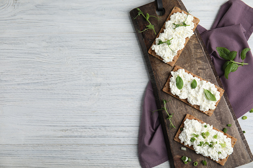 Crispy crackers with cottage cheese and different herbs on white wooden table, flat lay. Space for text