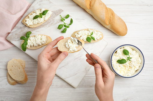 Woman spreading cottage cheese onto bread at white wooden table, top view