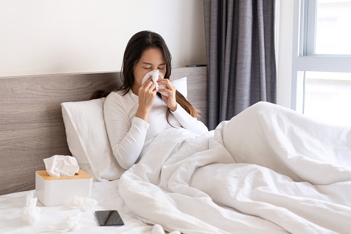 Sick young Asian woman feeling cold and has a high fever blowing nose while lying down on bed in bedroom at home. Winter cold and flu concept.