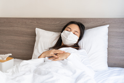 Asian woman has a high fever or infected with COVID-19 stay home and self quarantining during Coronavirus Pandemic. Ill girl wearing face mask have a headache and cough lying down on bed.