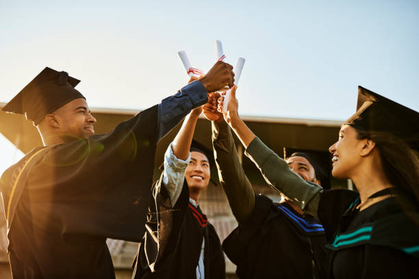 eine gruppe fröhlicher menschen bei der graduierung, die gemeinsam diplome oder zertifikate hochhalten und erfolge feiern. diverse multiethnische junge studenten in schwarzen gewändern außerhalb der universitätshochschule - graduation color image people photography stock-fotos und bilder