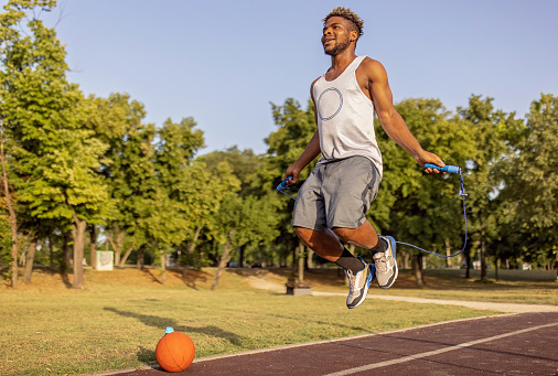 A young African American man wearing sports clothes is caught mid-air while jumping rope in a public park.