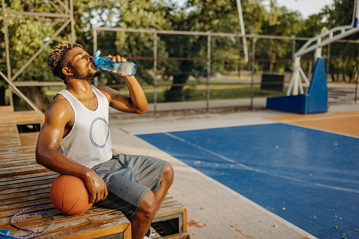 A young African American man is drinking from a water bottle and resting from his basketball practise.