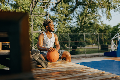 A young African American man is holding his hand over a basketball, while taking a rest on a bench.