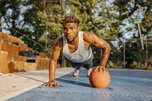 A young African American man in sports clothes is holding a push-up position, with his hand on a basketball.