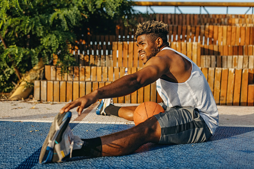 A young African American man is sitting on the floor and stretching after a basketball practise.