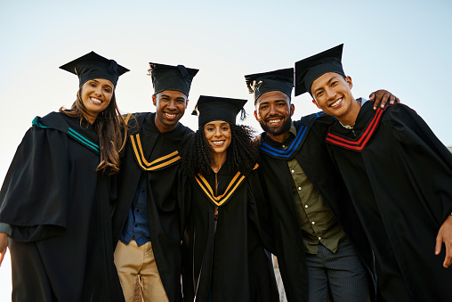 Portrait of diverse group of students in graduation gowns and caps with their arms around each other during graduation ceremony on university campus. Smiling friends standing close together at college