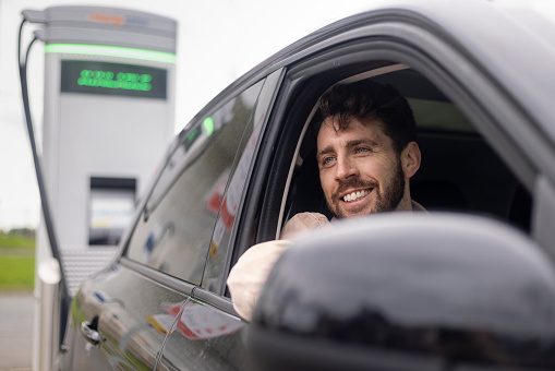 A young male adult smiling in his electric car. The Car is charged. He is smiling looking out of his car window wearing casual clothing.