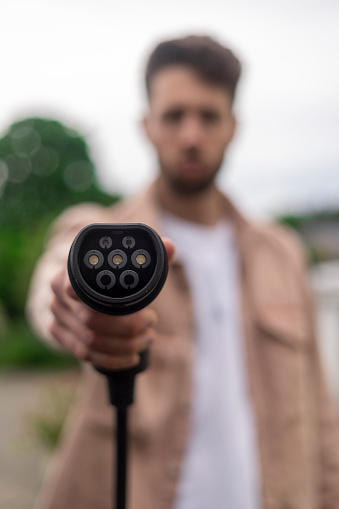 Unrecognisable young male adult holding up a car charger standing up outside wearing casual clothing.