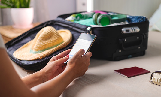 Close up of females hands packing suitcase for summer vacation. Checking the plane ticket online on the smart phone. Suitcase is on the bed  in bedroom.