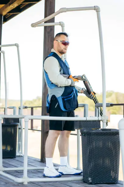 Photo of Adult man in sunglasses and a rifle vest practicing fire weapon shooting. Young experienced male aiming shotgun in outdoor.