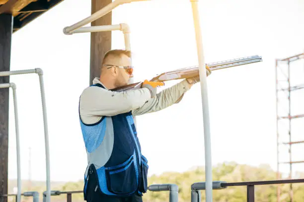 Photo of Adult man in sunglasses and a rifle vest practicing fire weapon shooting. Young experienced male aiming shotgun in outdoor.